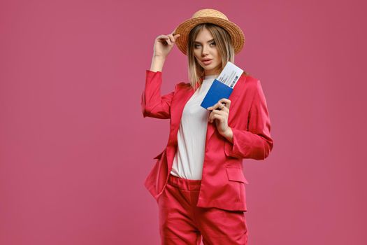 Alluring blonde girl in straw hat, white blouse and red pantsuit. She is touching her headdress, holding passport and ticket while posing on pink background. Travelling concept. Close-up, copy space
