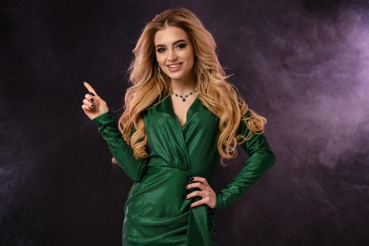 Gorgeous blonde girl, make-up, in green stylish dress, jewelry. Smiling, pointing at something, posing on black smoky background. Template, mockup for your advertising and design. Close up, copy space