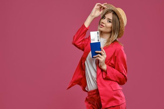 Alluring blonde lady in straw hat, white blouse and red pantsuit. Smiling, touching headdress, holding passport and ticket while posing on pink background. Travelling concept. Close-up, copy space