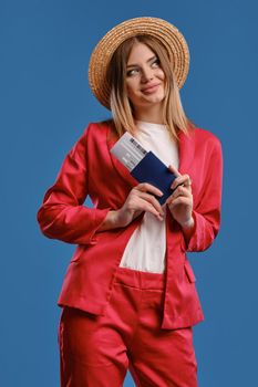 Gorgeous blonde girl in straw hat, white blouse and red pantsuit. She is smiling, looking aside, holding passport and ticket while posing on blue background. Travelling concept. Close-up, copy space
