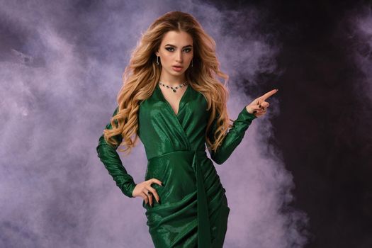 Attractive blonde female in green stylish dress, jewelry. Pointing at something by forefinger, posing on black smoky background. Template, mockup for your advertising and design. Close up, copy space