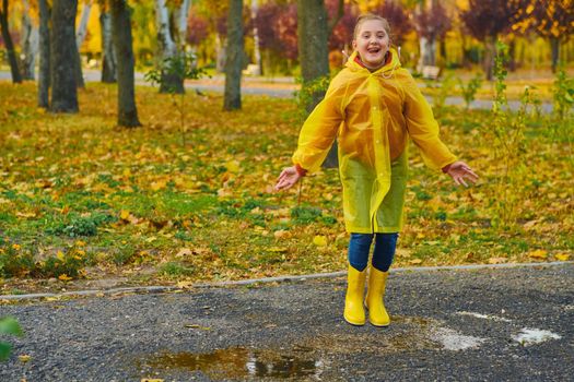 A happy girl in a yellow raincoat and rubber boots playing on an autumn walk. Kid with autumn leaves.