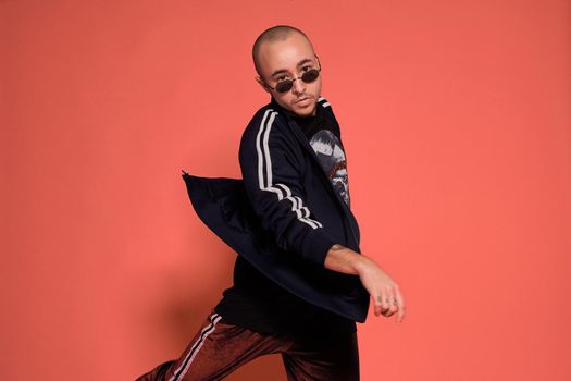 Close-up studio shot of a adult tattoed bald fellow in sunglasses, with a pirsing ring in his nose, wearing black trendy t-shirt with print and sport suit, fooling around while posing against a pink background with copy space. People, style and fashion concept. 90s style