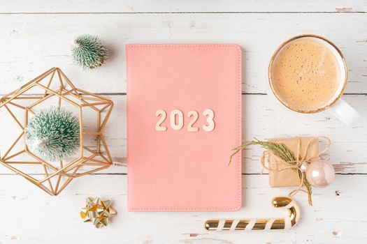 Christmas pink notepad with 2023 letters text. Flat lay on white wooden table background with planner, cup of coffee, candle, Christmas decoration, notebook and stationery. Top view Desktop