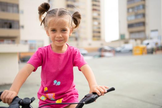 Cute little smiling girl riding bike bicycle in city on parking sunny summer day. Active family leisure with kids. Little caucasian girl 5 years old have fun with pink child bicycle