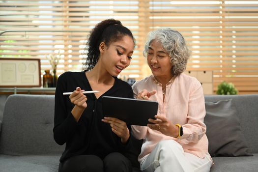 Smiling retried woman and her granddaughter surfing internet on digital tablet together on couch. Retirement, technology concept.