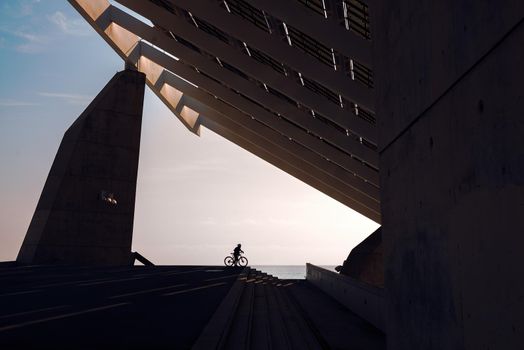 silhouette of a cyclist under a very famous steel and concrete structure of the Forum of Barcelona. The biker is next to some stairs and in the background you can see the sea, the sky is clear
