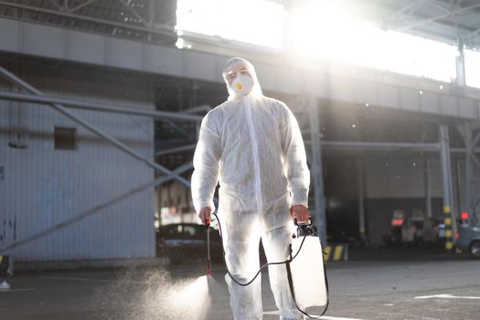 Man dressed white protective overalls spraying surface antibacterial sanitizer sprayer during quarantine Caucasian person protective suit disinfects parking during covid pandemic backlight