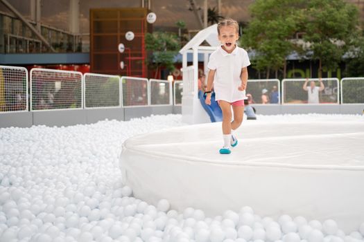 Happy little girl playing white plastic balls pool in amusement park. playground for kids. Active leisure for children.