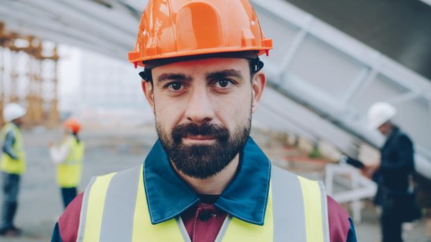 Portrait of young man engineer standing outdoors in workplace smiling looking at camera wearing uniform helmet and safety vest. People and occupation concept.
