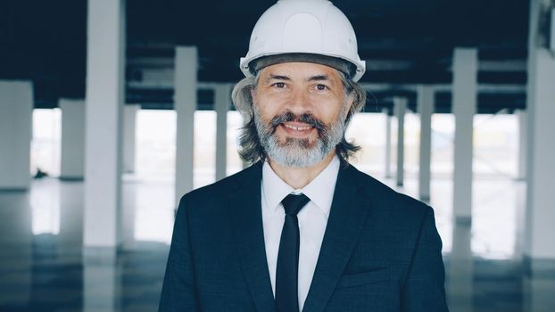 Portrait of successful real estate investor standing indoors in modern empty building wearing safety helmet and formal suit. People and occupation concept.