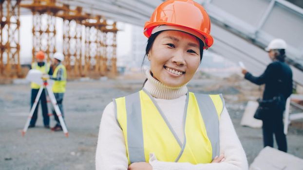 Cheerful Asian construction worker wearing uniform is smiling at construction site while people are working in background. Profession and emotions concept.