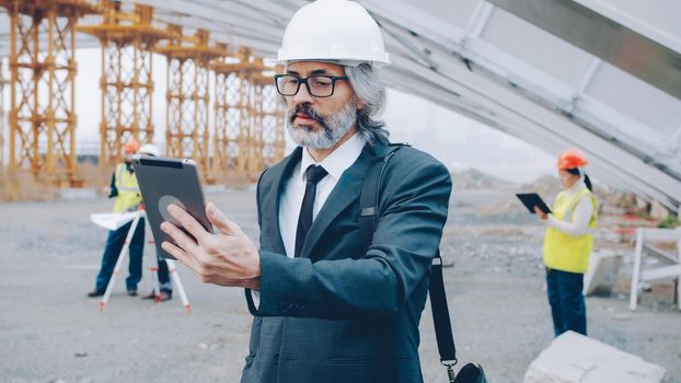Architect making online video call with tablet talking and looking at display standing at construction site while builders are working in background