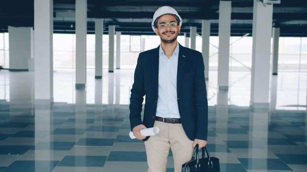 Portrait of young ambitious Middle Eastern engineer standing in new empty building holding blueprint and briefcase and looking at camera