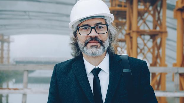 portrait of successful mature man architect wearing safety helmet standing in construction area alone and looking at camera