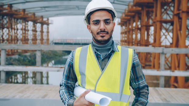 portrait of Arab construction specialist in helmet standing in building site holding plan and looking at camera. People and industry concept.