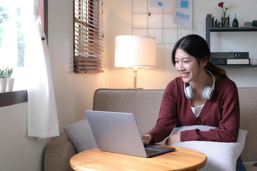 Beautiful Asian woman using laptop while headphones sitting on sofa at home.