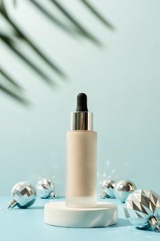 A minimalistic scene of foundation dropper bottle on podium with christmas decorative balls and pine tree on light blue background. Showcase with a stage for products, mockup design, seasonal