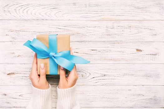 Woman hands give wrapped valentine or other holiday handmade present in paper with blue ribbon. Present box, decoration of gift on white wooden rustic vintage table, top view with copy space.