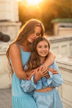 Portrait of mother and daughter in blue dresses with flowing long hair against the backdrop of sunset. The woman hugs and presses the girl to her. They are looking at the camera