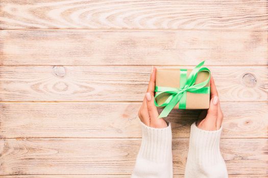 Woman hands give wrapped valentine or other holiday handmade present in paper with green ribbon. Present box, decoration of gift on wooden table, top view with copy space.