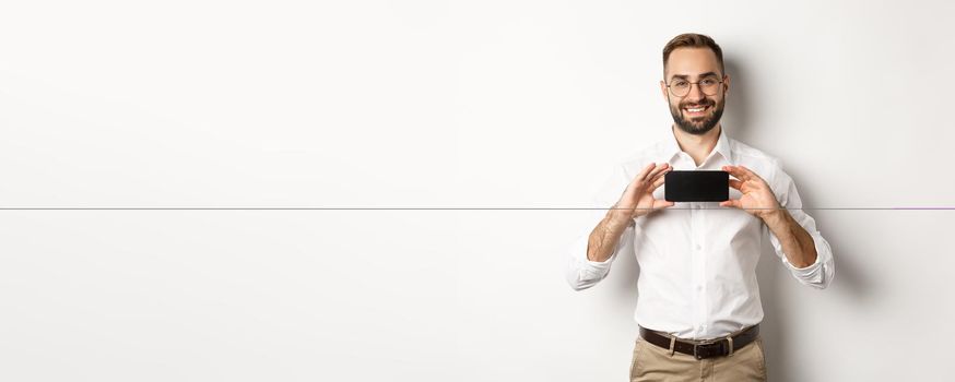 Handsome bearded man in glasses, demonstrating mobile phone application, showing smartphone screen, standing over white background.