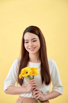 Beautiful woman in the white shirt with flowers gerbera in hands on a yellow background. She smiles and laughs