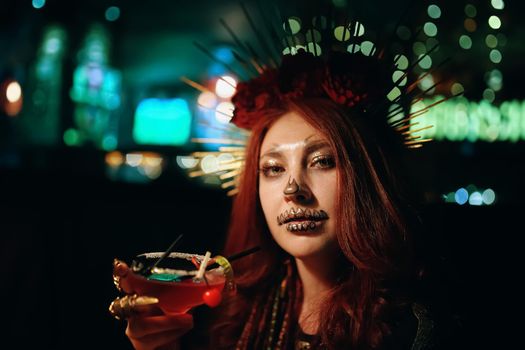 Female with skeleton makeup at a Halloween party holds cocktail and looks at camera. All Saints' Night. Celebration in a nightclub. Woman in creepy costume of Mexican goddess of death.