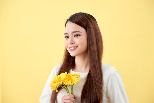 Beautiful woman in the white shirt with flowers gerbera in hands on a yellow background. She smiles and laughs