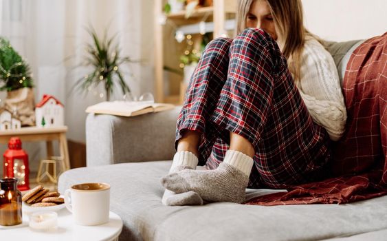 Cozy woman legs in knitted winter warm socks, sweater and checkered plaid drinking hot cocoa or coffee in mug, during resting on couch at home. Christmas holidays with candle, decor and cookies