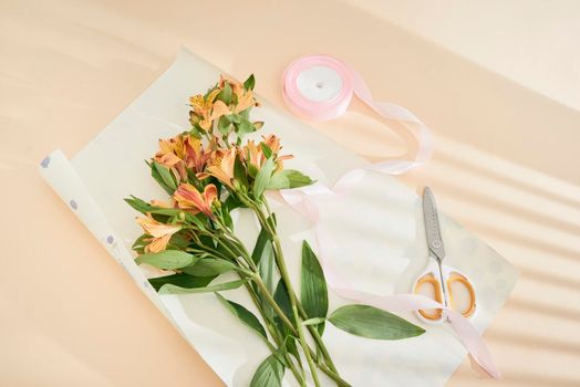 top view of beautiful orange flowers, scissors, ribbon and craft paper on paper surface