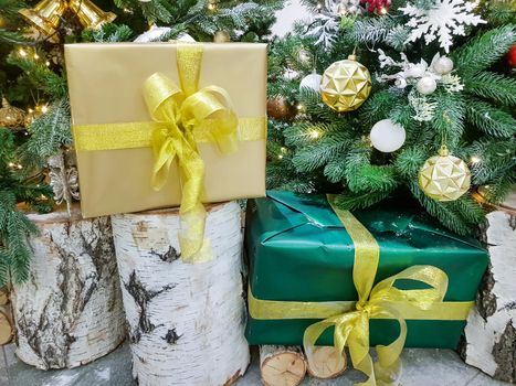 Beautiful boxes wrapped in gold paper with Christmas gifts on a birch stump near the Christmas tree in the room.