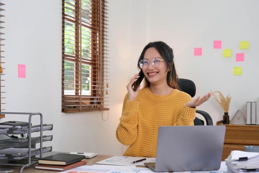 Attractive and smiling Asian businesswoman is talking on a mobile phone with a customer with laptop in the office..