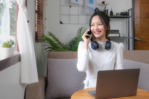 Charming young Asian female relaxes in the living room enjoying talking with her coworker on her phone while working from home..