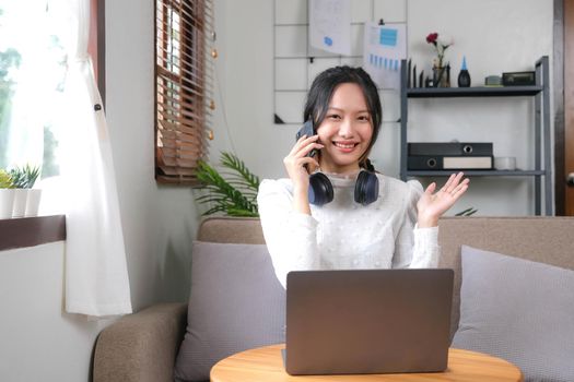 Happy and cheerful young Asian female relaxes in the living room enjoying talking with her friends on a video call through her smartphone..