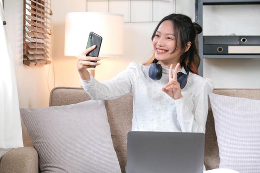 Happy and cheerful young Asian female relaxes in the living room enjoying talking with her friends on a video call through her smartphone..