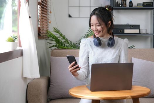 Happy and cheerful young Asian female choosing her music playlist on mobile phone, listening to music through her headphones while relaxing in the living room..