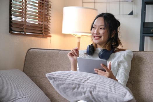 Attractive young Asian female with wireless headphones relaxes in the minimal living room using a digital tablet..