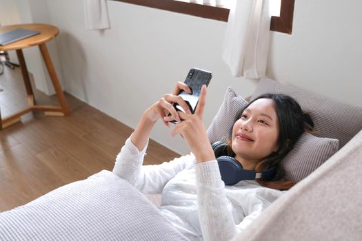 Female lying leaning on comfortable sofa and using her modern mobile phone to chat with her friends, scroll on shopping application or social media