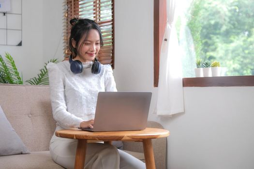 Portrait of young female with headphone using laptop while relaxed sitting on sofa.
