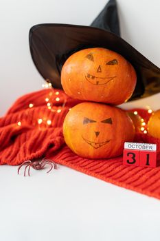 Two pumpkins with a painted face in a hat, a spider, a wooden calendar on a red sweater. Holiday concept of October 31, Halloween. Beautiful bokeh on the background