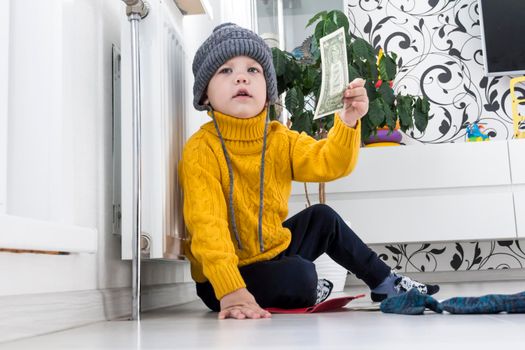 A little boy in a yellow sweater and hat is counting money and studying heating bills, near a heater with a thermostat..