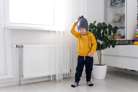 A small child in a yellow sweater and a gray hat is standing near a heater with a thermostat.,,