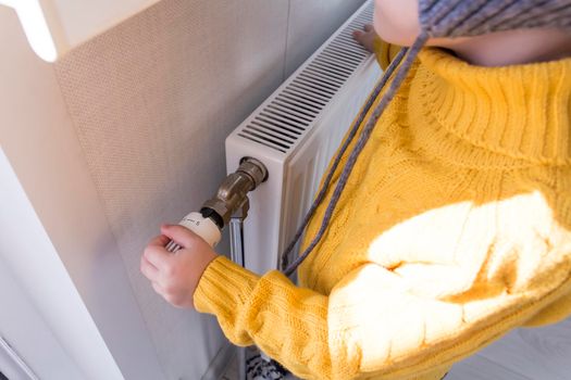 A small child in a yellow sweater and a gray hat is standing near a heater with a thermostat.,,