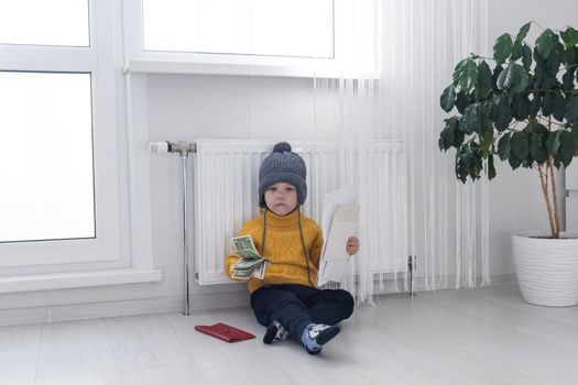 A little boy in a yellow sweater and hat is counting money and studying heating bills, near a heater with a thermostat..