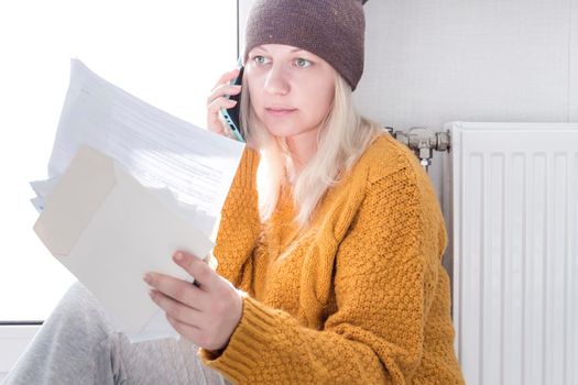 A young girl in a yellow sweater and a brown hat is sitting on the floor, counting money and thinking how to pay bills and taxes near a heater with a thermostat..