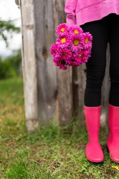 A young girl in pink rubber boots stands against the background of an old house in the garden and holds a bouquet of pink flowers in her hands.