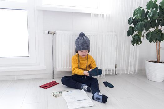 A little boy in a yellow sweater and hat is counting money and studying heating bills, near a heater with a thermostat..