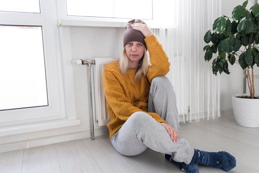 A young girl in a hat and a yellow sweater is sitting on the floor and holding her head near a heater with a thermostat..