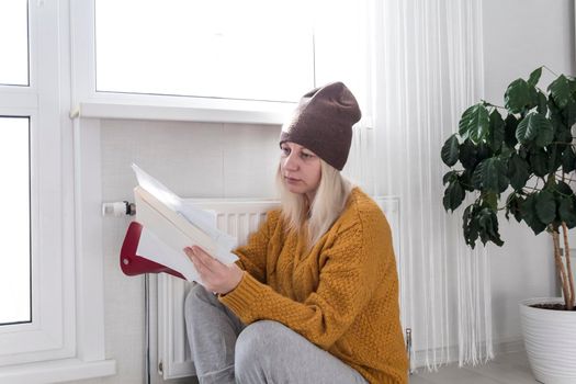 A young girl in a yellow sweater and a brown hat is sitting on the floor, counting money and thinking how to pay bills and taxes near a heater with a thermostat..